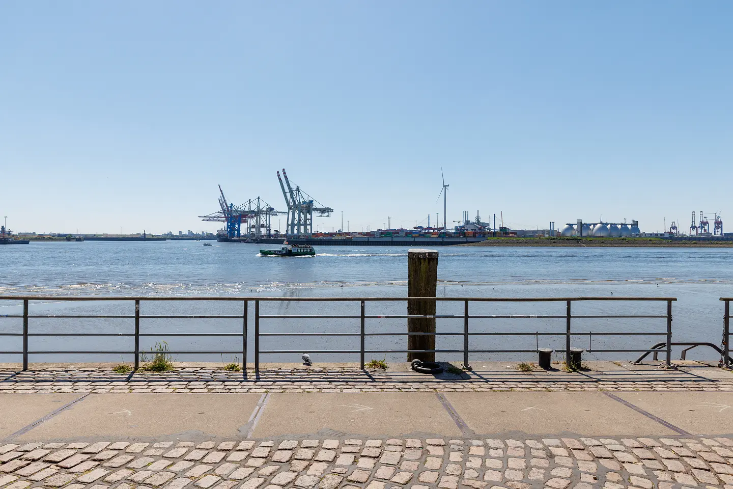 Blick auf den Hamburger Hafen mit Containerschiffen, Hafenkränen und Windkraftanlage bei sonnigem Wetter – aufgenommen vom Elbufer aus, ganz in der Nähe der Kanzlei von Rechtsanwalt Dr. Mackenzie-Owen